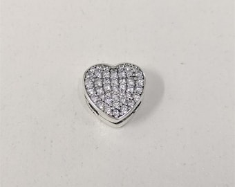 May include: A silver heart-shaped charm, encrusted with numerous small, clear gemstones. The charm has a polished, reflective surface and is designed with a hinged opening. The gemstones cover the front of the heart.