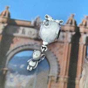 Può includere: Un charm in argento raffigurante una piccola figura a forma di orso che tiene in mano un charm a forma di palloncino. Il palloncino è bianco con un effetto marmorizzato e ritagli a forma di cuore. Il charm è progettato per un braccialetto o una collana.