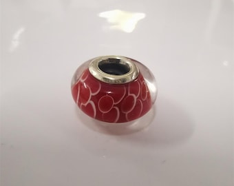 May include: A red and white glass bead with a silver-colored metal rim. The bead has a pattern of white ovals on a red background. The bead is clear and transparent.