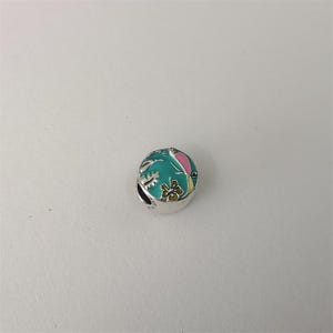 Puede incluir: Un charm de plata con un diseño de esmalte turquesa que presenta un pájaro rosa y amarillo, una flor y follaje. El charm es redondo con un pequeño agujero en el lateral.