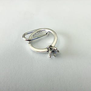 Può includere: Un anello d'argento con una gemma trasparente a taglio quadrato. L'anello ha un design unico con un anello che incrocia la fascia principale. Le parole "Self Love" sono incise sull'anello.