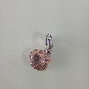 May include: A rose gold seashell charm with a silver loop. The charm has a textured surface and a small, sparkling accent. The seashell design is detailed and elegant, perfect for a bracelet or necklace.