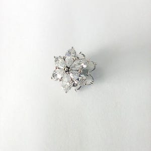 May include: A silver-toned flower-shaped jewelry piece with clear, teardrop-shaped stones. The stones are arranged in a floral pattern, with each stone pointing outwards from the center. The piece is set against a white background.