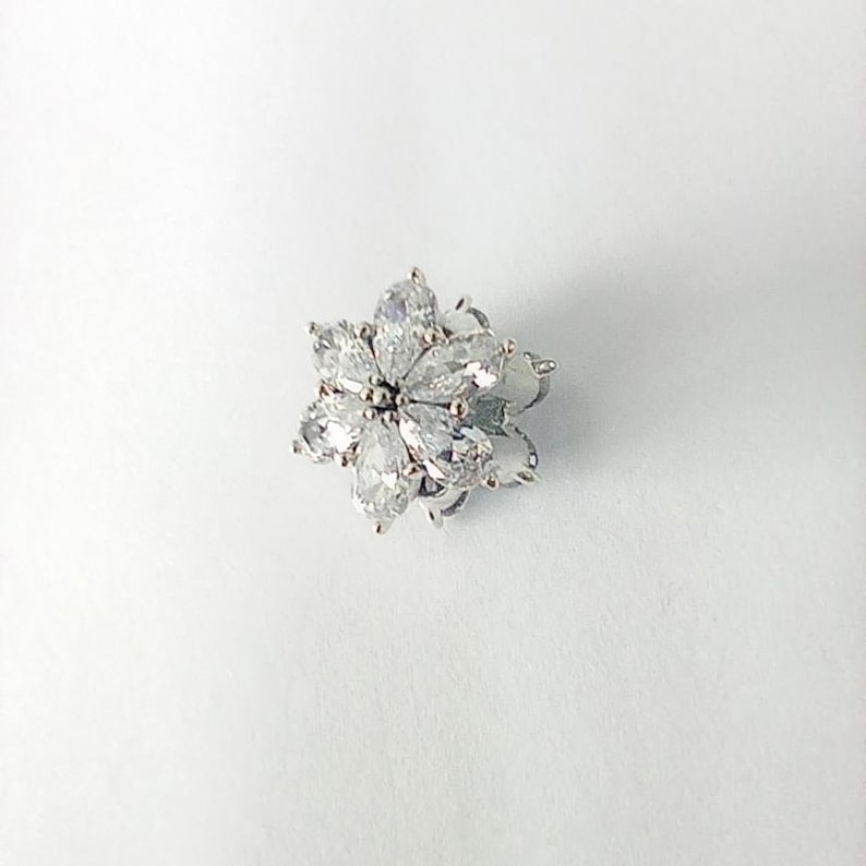May include: A silver-toned flower-shaped jewelry piece with clear, teardrop-shaped stones. The stones are arranged in a floral pattern, with each stone pointing outwards from the center. The piece is set against a white background.