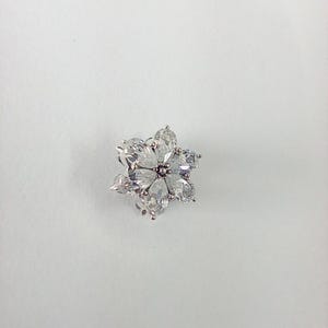 May include: A flower-shaped brooch with clear, pear-cut gemstones. The brooch features six petals surrounding a central gemstone, all set in a silver-toned metal. The design is elegant and suitable for formal wear.