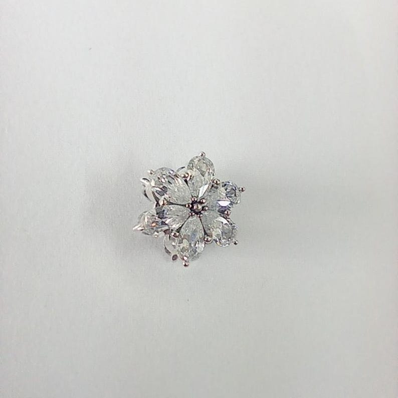 May include: A flower-shaped brooch with clear, pear-cut gemstones. The brooch features six petals surrounding a central gemstone, all set in a silver-toned metal. The design is elegant and suitable for formal wear.