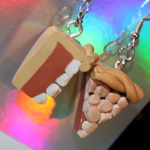 May include: A pair of handmade earrings featuring miniature pie slice charms. One earring depicts a slice with a red filling and white accents, while the other shows a pie with a tan crust and small, round details. The earrings hang from silver-colored hooks.