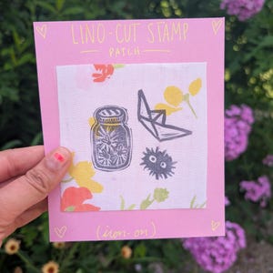 May include: A pink card with the text "LINO-CUT STAMP PATCH" and a fabric patch with linocut designs of a jar, origami crane, and a round creature. The patch is decorated with floral patterns and the text "(iron-on)".
