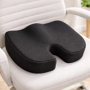 May include: A black ergonomic seat cushion with a contoured design, placed on a white office chair. The cushion has a U-shaped cutout in the front and a textured surface, designed for comfort and support.