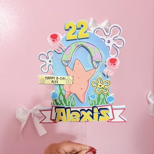 Peut inclure: Une décoration de gâteau en papier superposé représentant un personnage d'étoile de mer, le nombre 22 et le prénom Alexis. Le design inclut des méduses, des algues et une bannière avec le texte HAPPY B-DAY ALEX, dans des tons bleu, rose et jaune.