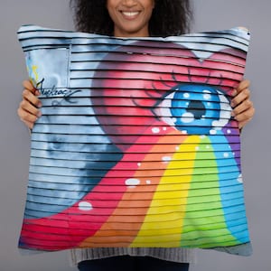 Peut inclure: Coussin carré avec un design graffiti vibrant. L'œuvre d'art comprend un grand œil bleu, un cœur rouge et un arc-en-ciel. Le fond présente un motif rayé bleu et gris, avec le mot "Muckroc" visible.