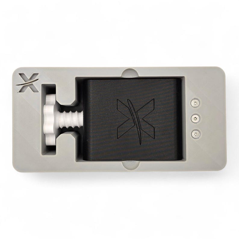 Peut inclure: Un cadre de bo&icirc;tier gris rectangulaire contenant un bloc central noir avec un logo X en relief. Un bouton de r&eacute;glage filet&eacute; blanc est positionn&eacute; sur la gauche, et trois petites fixations m&eacute;talliques circulaires sont align&eacute;es sur la droite.