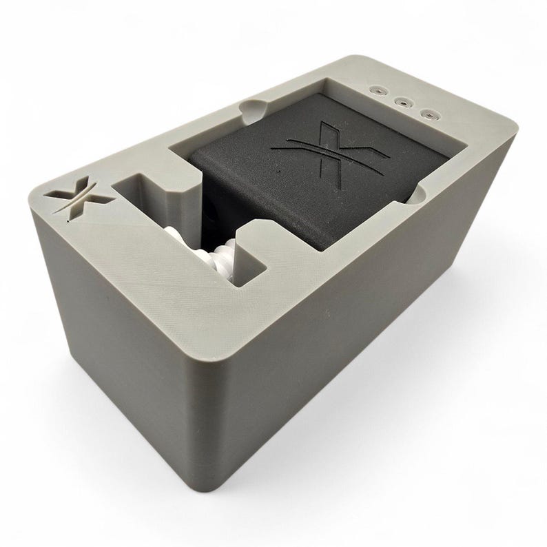 Peut inclure: Une station d'accueil rectangulaire grise au fini mat, dot&eacute;e d'un composant carr&eacute; noir encastr&eacute; avec un logo X stylis&eacute; en relief sur le dessus. La base grise pr&eacute;sente &eacute;galement un logo X en relief et trois petits ports circulaires sur le c&ocirc;t&eacute;.