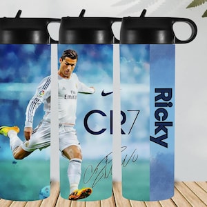 Pode incluir: Uma garrafa de água preta com um jogador de futebol de uniforme branco a chutar uma bola. A garrafa tem uma tampa preta e um fundo azul e branco. O texto "CR7" e uma assinatura são visíveis.