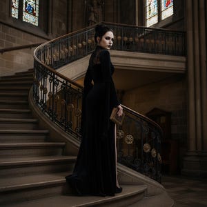 May include: A long black velvet gown with bell sleeves and a high slit. The dress features a lace panel on the back and is worn on a grand, curved staircase with ornate iron railings. The person is holding a small, antique book.