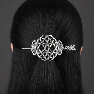 Op de afbeelding: Een zilverkleurige haarspeld met een ingewikkeld, cirkelvormig Keltisch knoopontwerp. Het accessoire wordt vastgezet met een bijpassende metalen pin die door het midden steekt, afgewerkt met een decoratief, gestructureerd uiteinde in pijl.