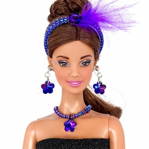 May include: A doll wearing a black strapless dress and a matching jewelry set. The set includes a flower pendant necklace, flower earrings, and a jeweled headband with a purple feather. The doll has brown hair and is wearing makeup.