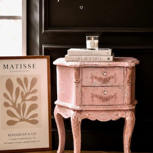 May include: A light pink wooden nightstand with two drawers, featuring ornate carved floral details and clear crystal-style knobs. The piece stands on four curved legs and is topped with two stacked books and a lit candle in a glass jar.
