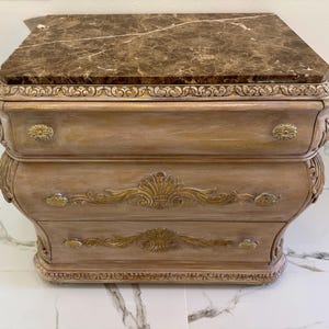 Puede incluir: Una mesita de noche de dos cajones con tapa de mármol y detalles dorados. La mesita tiene un acabado marrón claro con tallas decorativas y herrajes dorados. La tapa de mármol tiene vetas marrones y beige.