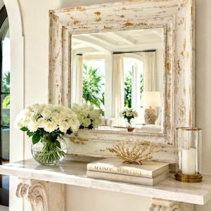 May include: A square wall mirror with a distressed white and gold frame featuring a beaded inner edge. The mirror reflects a bright room with white curtains and a lamp. The frame has a weathered finish with visible gold leaf accents.