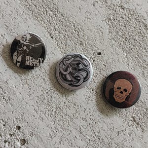 Puede incluir: Tres chapas redondas. La chapa de la izquierda es en blanco y negro con una figura y el texto HEAVY METAL. La chapa central es blanca con una ilustración negra de una serpiente enroscada. La de la derecha muestra una calavera marrón.