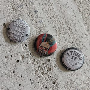 Puede incluir: Tres chapas redondas. La chapa de la izquierda es plateada con el texto FUCK IT LETS RIDE. La chapa central muestra una ilustración colorida de una calavera con alas. La chapa de la derecha es plateada con el texto I TRIED IT LIKED IT.