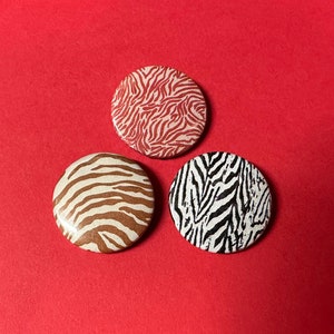 May include: Three round buttons with different animal print designs. One button has a red and white zebra print, one has a brown and white zebra print, and one has a black and white zebra print.