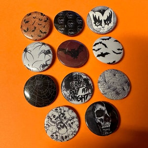 Puede incluir: Diez pines de botón temáticos de Halloween en blanco y negro. Los pines presentan varios diseños que incluyen murciélagos, arañas, calaveras y una telaraña. Un pin presenta el texto "We Come Out At Night".