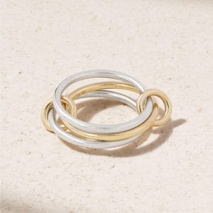 May include: A mixed metal ring featuring interlocking circles. The ring combines silver and gold tones, with three larger silver bands and two smaller gold bands. The design is modern and geometric, suitable for everyday wear.