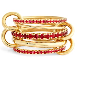 May include: A gold-colored ring set with multiple bands linked together. Two of the bands are encrusted with small, vibrant red gemstones. The ring has a modern, elegant design, perfect for adding a touch of luxury to any outfit.