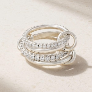 May include: A silver ring set featuring multiple interlocking bands. Two bands are encrusted with small, clear gemstones. The rings are interlocked, creating a layered effect. The jewelry is displayed on a light-colored surface.