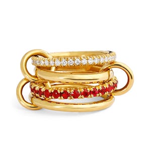 Op de afbeelding: Een stapel van vier gouden ringen. De bovenste ring is bezet met kleine, heldere stenen. De tweede ring is effen goud. De derde ring is bezet met kleine, rode stenen. De onderste ring is effen goud. De ringen zijn met elkaar verbonden.