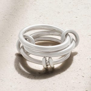May include: A silver ring with an unusual design. The ring has several interlocking bands, giving a layered appearance. The metal has a matte finish, and the ring is set against a neutral background.