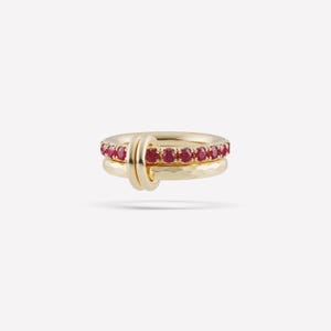 May include: Three gold rings. The top ring is adorned with small, red gemstones. The middle ring is connected to the top ring with a gold loop. The bottom ring has a hammered texture. The rings are set against a white background.