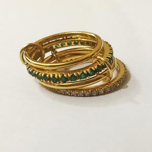 Może przedstawiać: Stos złotych pierścionków. Jeden pierścionek jest inkrustowany małymi zielonymi kamieniami szlachetnymi, drugi przezroczystymi kamieniami szlachetnymi, a pozostałe są gładkie. Pierścionki są ułożone w zagnieżdżonej konfiguracji, tworząc warstwowy efekt.