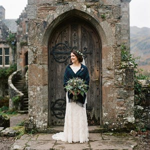May include: A floor-length white lace wedding dress with a sweetheart neckline, paired with a dark green and navy blue plaid wool shawl with fringe edges. The ensemble includes a floral headpiece and a bouquet of greenery and thistle flowers.