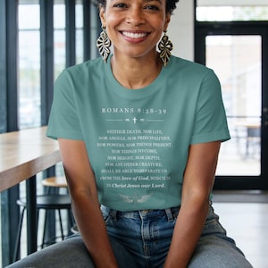 Könnte beinhalten: Ein kurzärmeliges T-Shirt in Blaugrün mit weißem Text, der Romans 8:38-39 lautet, gefolgt vom vollständigen Bibelvers und einer kleinen Grafik eines Kreuzes mit Flügeln am unteren Rand. Das T-Shirt wird mit einer blauen Jeans kombiniert.