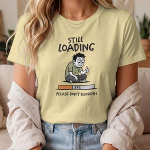 May include: A pale yellow t-shirt featuring a graphic of a tired person sitting with a thumbs up. Above the figure is the text STILL LOADING, and below is a progress bar at 37% with the text PLEASE DON'T REFRESH written underneath.
