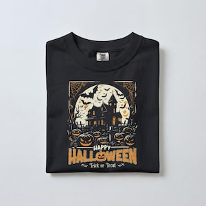 A black t-shirt neatly folded on a plain white surface, displaying a haunted house and jack-o-lantern graphic with Halloween text.