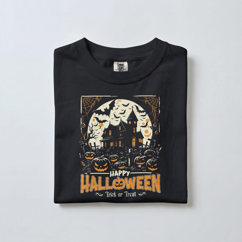 A black t-shirt neatly folded on a plain white surface, displaying a haunted house and jack-o-lantern graphic with Halloween text.