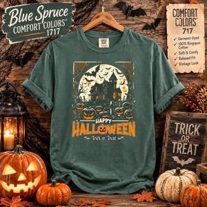 A blue spruce green t-shirt laid flat on wood with a haunted house and pumpkin graphic, accompanied by decorative pumpkins and a lantern.