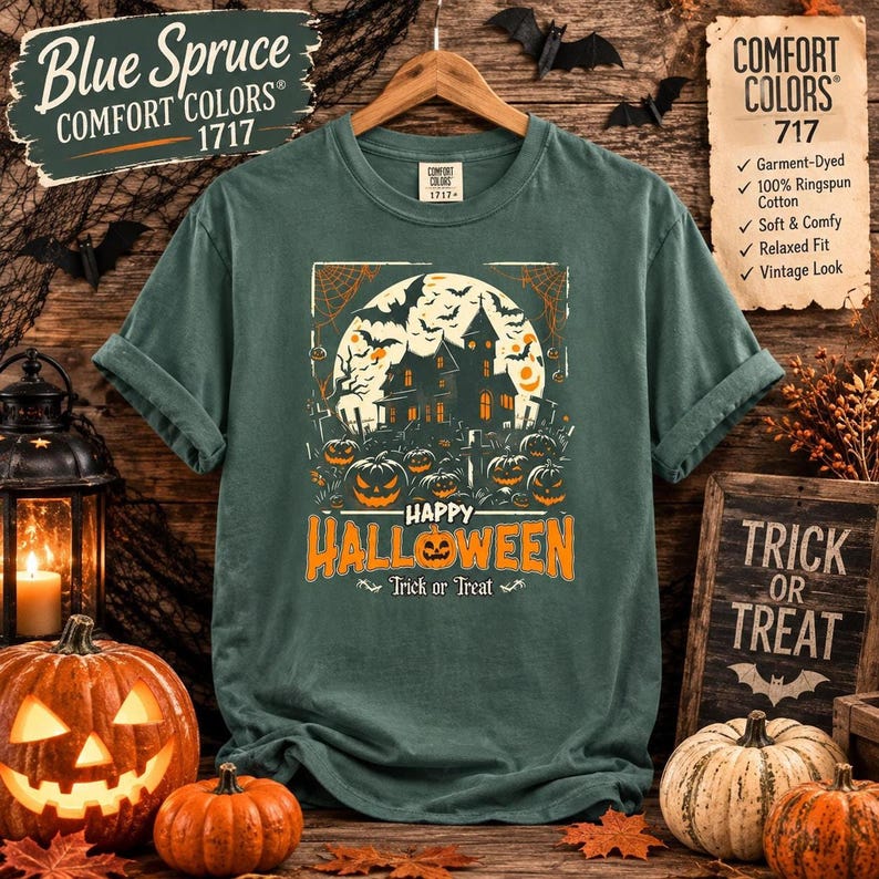 A blue spruce green t-shirt laid flat on wood with a haunted house and pumpkin graphic, accompanied by decorative pumpkins and a lantern.