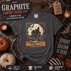 A graphite gray t-shirt laid flat on a wooden surface, featuring a haunted house and pumpkin graphic alongside Halloween decor.