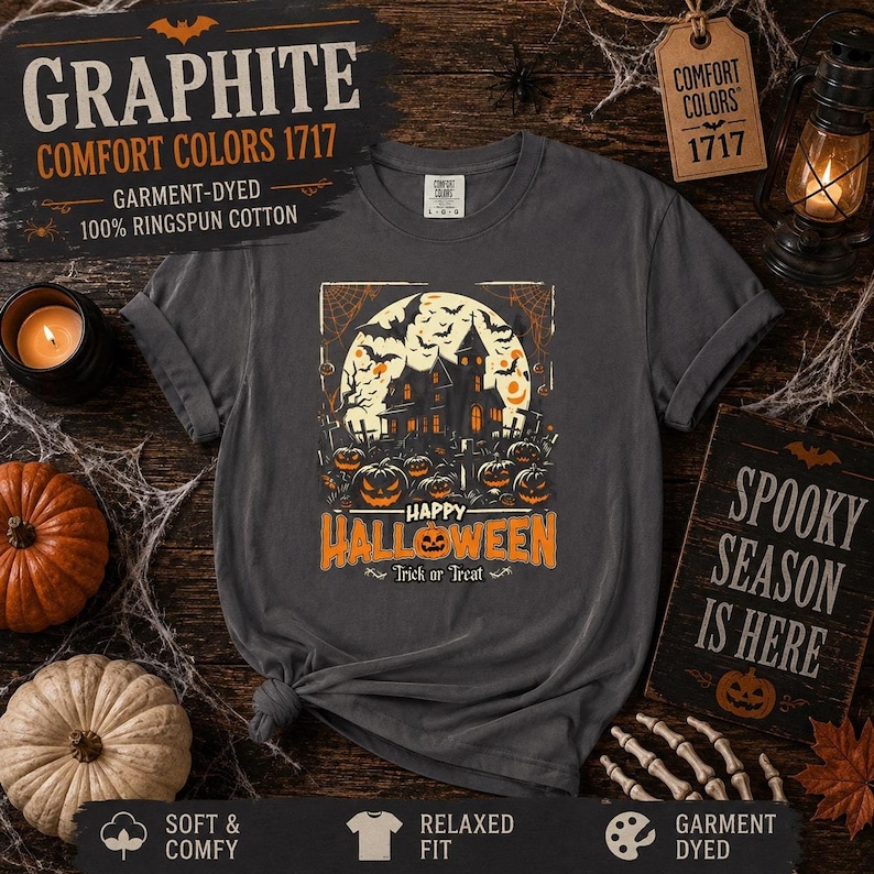 A graphite gray t-shirt laid flat on a wooden surface, featuring a haunted house and pumpkin graphic alongside Halloween decor.
