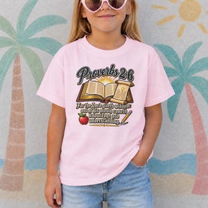Könnte beinhalten: Ein hellrosa T-Shirt mit kurzen Ärmeln und einem Grafikdruck, der ein offenes Buch, eine Schriftrolle, einen roten Apfel und einen Bleistift zeigt. Der Text lautet Proverbs 2:6, For the Lord giveth wisdom: out of his mouth cometh knowledge and understanding.