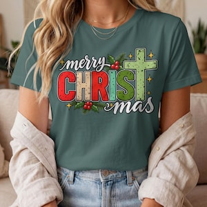 May include: A dark sage green short-sleeve t-shirt featuring a graphic print that reads merry CHRISTmas with a cross replacing the letter T. The design includes red and green lettering, holly berry accents, and small yellow star shapes.