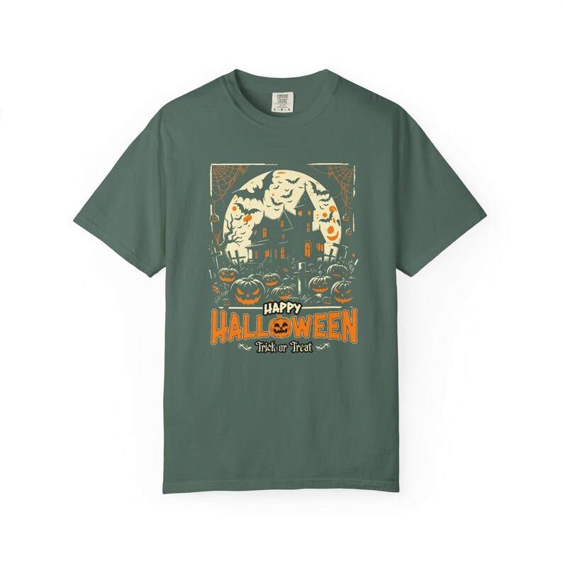 May include: A dark sage green t-shirt featuring a graphic print of a haunted house silhouette against a large full moon, surrounded by bats, spiderwebs, and jack-o'-lanterns. The text reads "HAPPY HALLOWEEN" and "Trick or Treat".