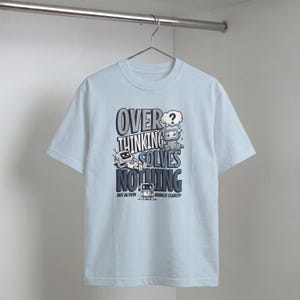 May include: Light blue t-shirt with the text "OVERTHINKING SOLVES NOTHING" and robot illustrations. The shirt also includes the phrase "BUT ACTION BRINGS CLARITY". The design is in shades of gray and white.