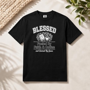 May include: A black short-sleeve t-shirt featuring white screen-printed text that reads BLESSED, Fueled By Faith & Coffee, and Covered By Grace. The design includes a graphic of a book with a cross on the cover and a steaming coffee cup.