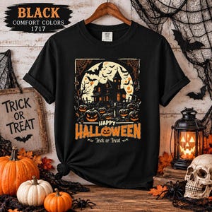 A black t-shirt laid flat on a wooden surface with a haunted house and pumpkin graphic, surrounded by Halloween props including pumpkins and a skull.